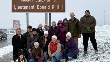 Group of people in front of brown ceremonial NYSDOT signs denoting "Lieutenant Donald R Hill Memorial Highway"