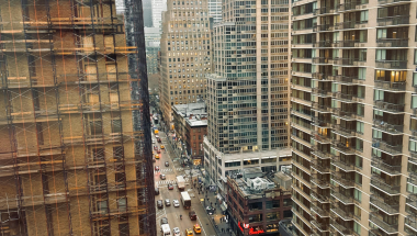 A high-angle view looking down a New York City street canyon on an overcast, foggy day. On the left, a historic brick building is covered in construction scaffolding. On the right, a modern high-rise residential building with balconies. The street below is active with yellow taxis, pedestrians, and a Chick-fil-A visible at street level. Skyscrapers disappear into the fog in the background.