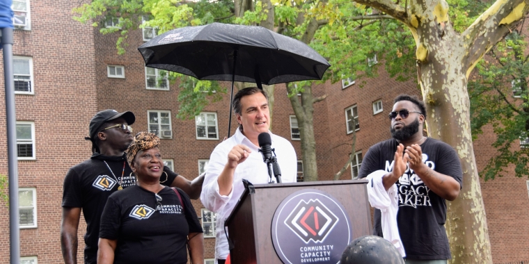 Senator Gianaris delivers remarks from a podium bearing the red-and-black logo of Community Capacity Development. Standing next to him are volunteers from the organization.