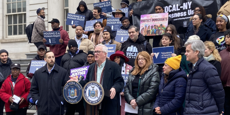 Alt text: Senator Brian Kavanagh speaks at a podium outside New York City Hall during a rally marking the launch of the Housing Access Voucher Program (HAVP). Standing with him are Assemblymember Linda Rosenthal, Assemblymember Jessica González-Rojas, Manhattan Borough President Brad Hoylman-Sigal, and Christopher Mann of Win. Housing advocates and community members stand on the steps behind them holding signs reading “Housing Is a Human Right” and banners from organizations including VOCAL-NY and Volunteer