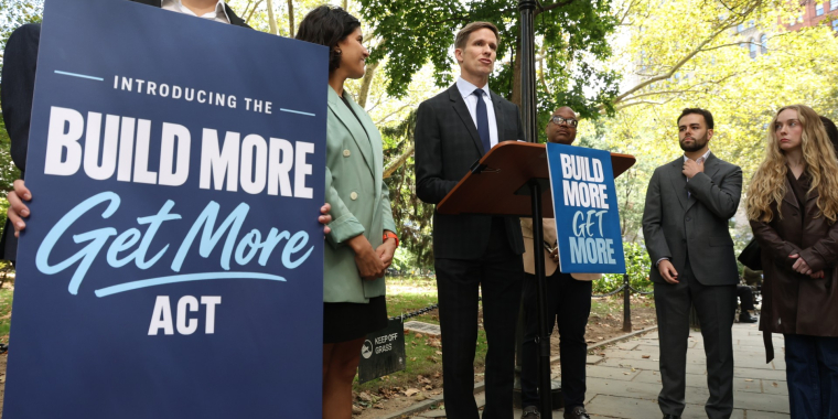 Senator Erik Bottcher stands behind a podium at a press conference in City Hall Park, with a sign displaying the name "Build More, Get More Act"