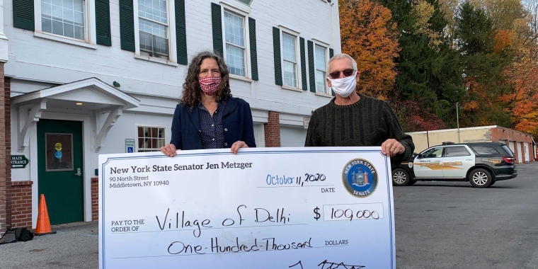 Senator Jen Metzger presents a giant check for $100K to Village of Delhi Mayor Richard Maxey in front of Delhi Village Hall on Sunday.